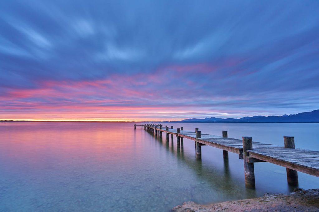 Dramatic sunrise colors over Chiemsee Lake with a wooden pier stretching into the calm water.